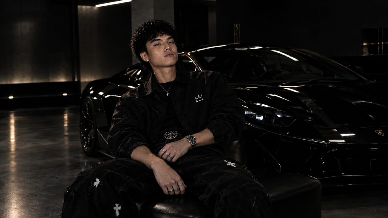 Kimchi in black seated beside a glossy black sports car in a dark garage